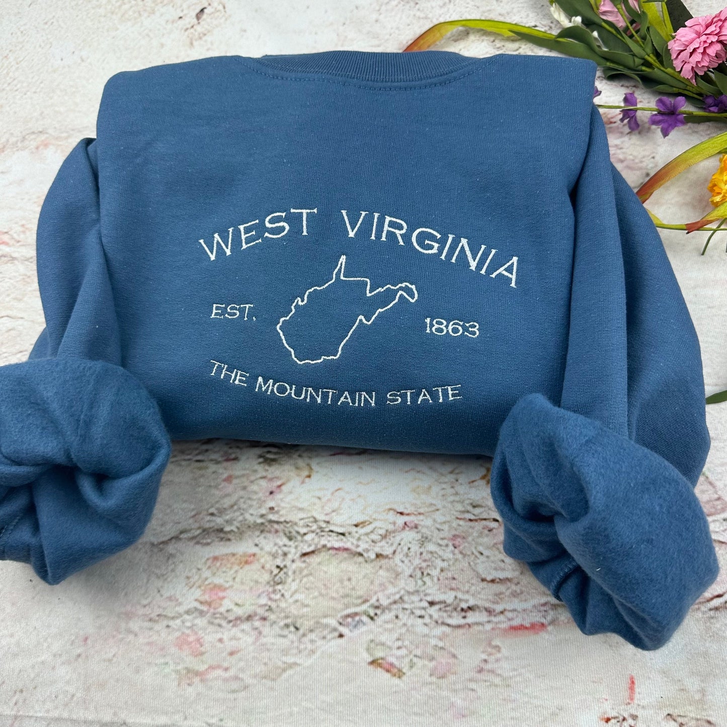 A blue sweatshirt with a white embroidered design. The design features the outline of the state of West Virginia, the words 'West Virginia', 'Est. 1863', and 'The Mountain State'.