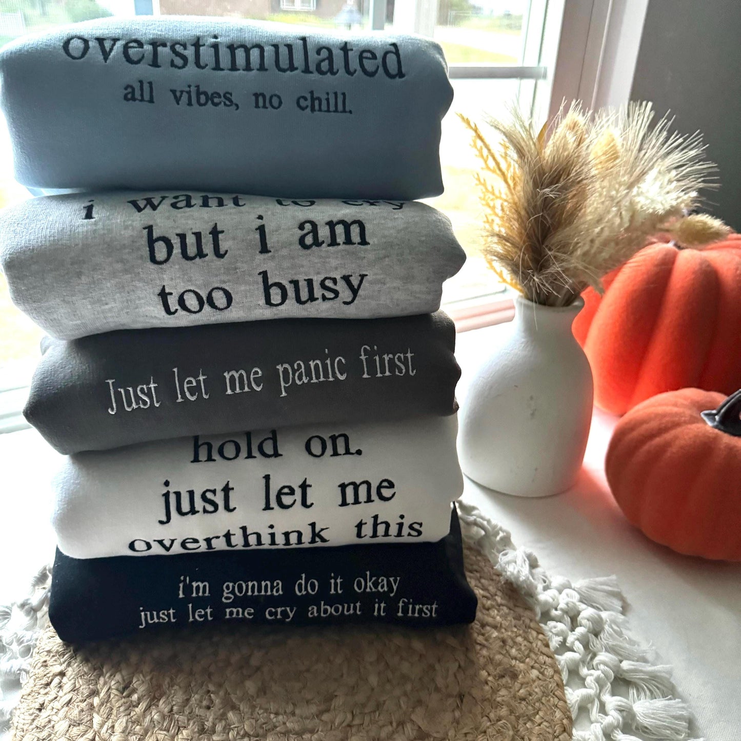 an image of 5 sweatshirt folded on top of one another. Each sweatshirt is a different color and have different text embroidered onto it in various thread colors.