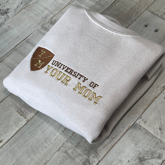 Unisex "University of Your Mom" embroidered sweatshirt in sand color, folded on wooden surface.