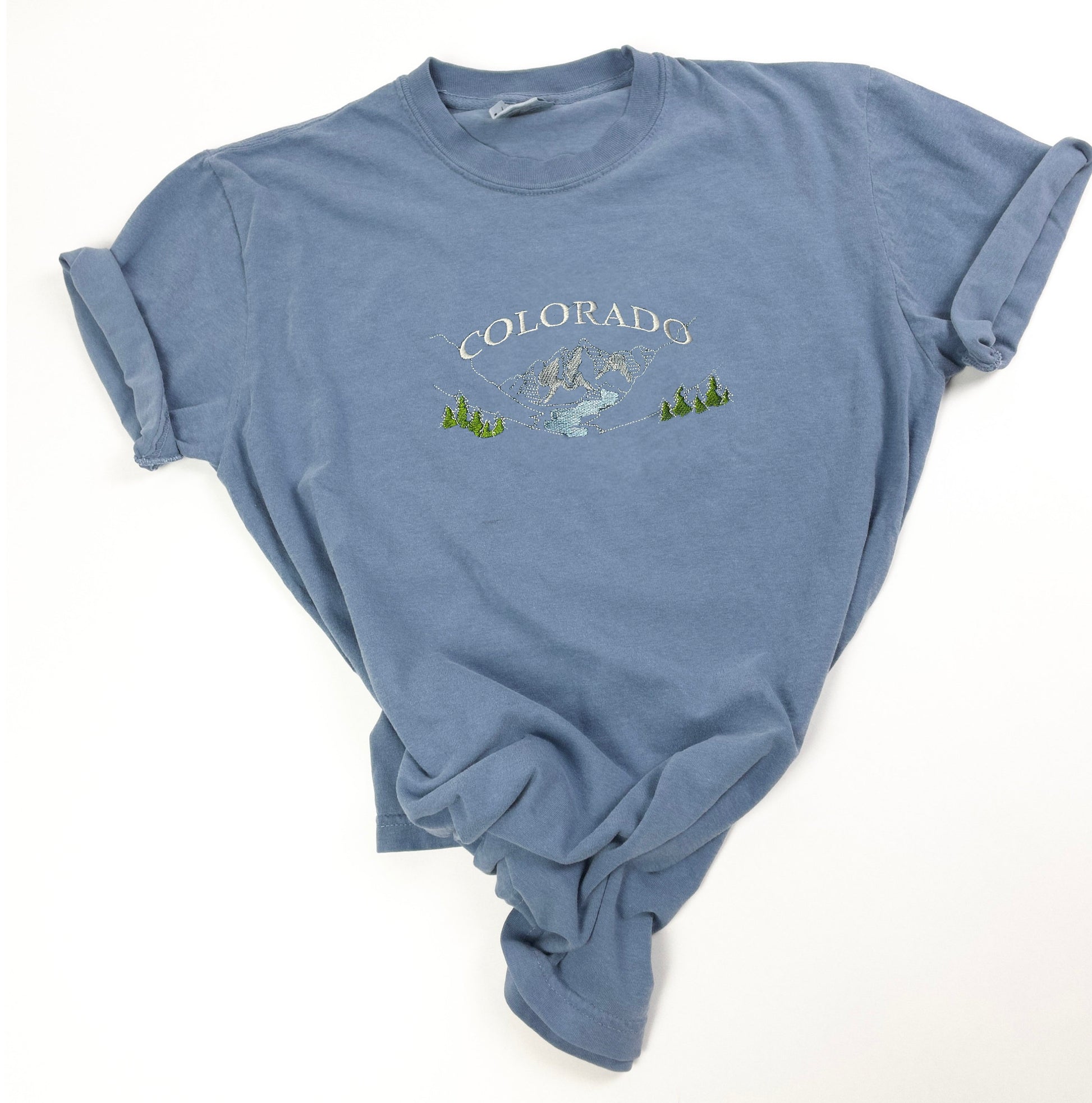A blue t-shirt with a white embroidered design of mountains and trees. The text &#39;COLORADO&#39; is embroidered in white above the mountains.