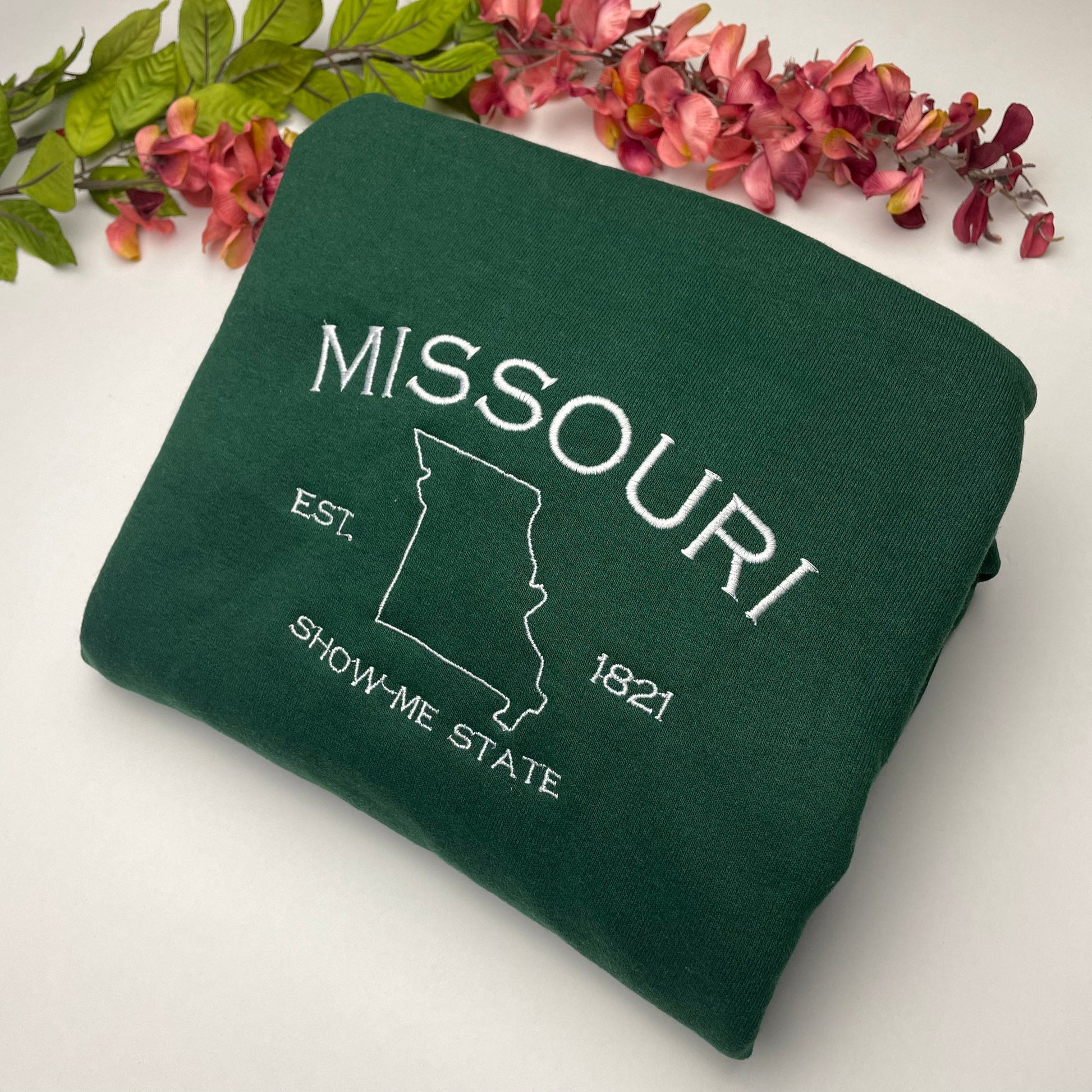 an image of a forest green sweatshirt with the words Missouri embroidered onto it. There is also an outline of the state of Missouri and the word show me state underneath that.