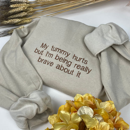 A beige colored sweatshirt with the words “My tummy hurts but I’m being really brave about it” is embroidered in brown thread