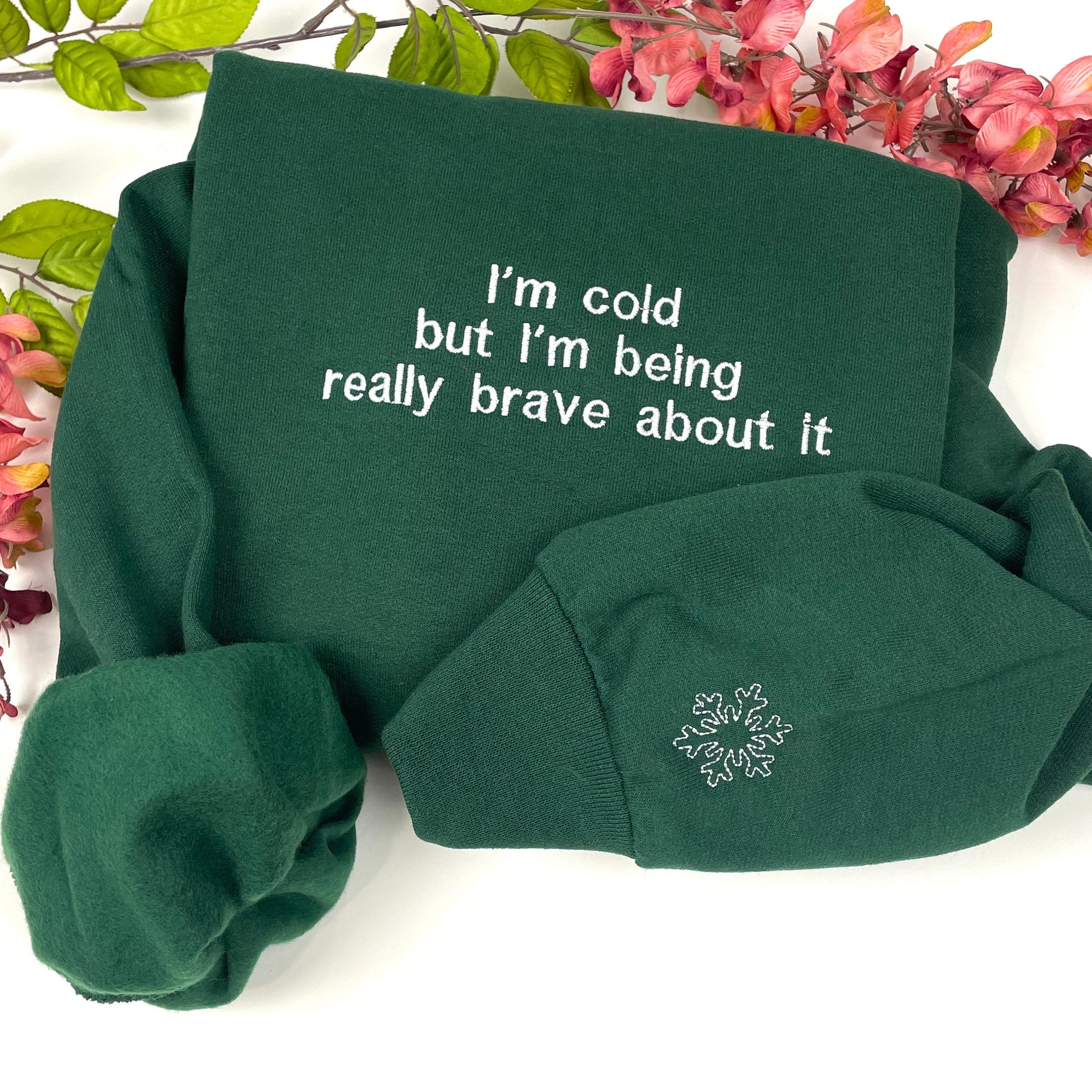 Green sweatshirt with "I'm cold but I'm being really brave about it" embroidery, folded with floral background.