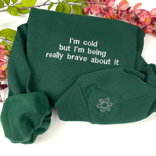 Green sweatshirt with "I'm cold but I'm being really brave about it" embroidery, folded with floral background.