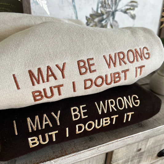 an image of two sweatshirts folded on top of one another. the top shirt is beige with brown lettering, the bottom shirt it brown with beige lettering. The words on the shirt in capital letters say &quot;I may be wrong but i doubt it&quot; in