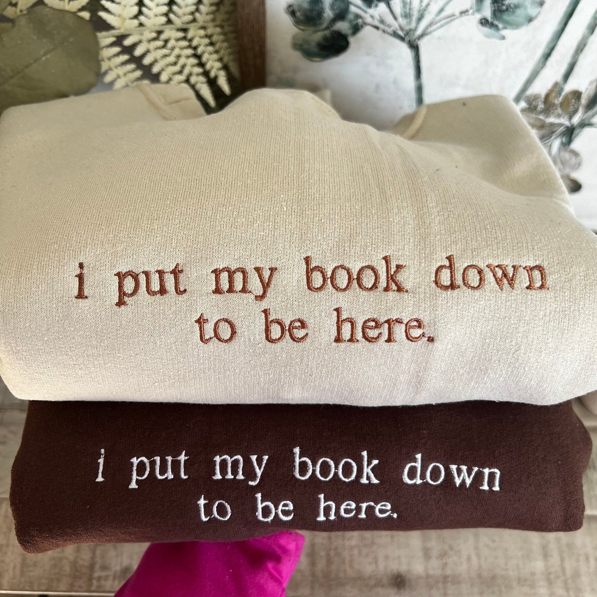 I picture of 2 sweatshirts folded on top of one another. The top sweatshirt is beige/sand and the bottom sweatshirt is brown. Both shirts have the words &quot;I put my book down to be here.&quot; embroidered onto them.