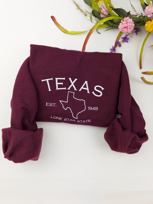 Texas embroidered sweatshirt with "Lone Star State" design in maroon, rolled sleeves, floral backdrop.