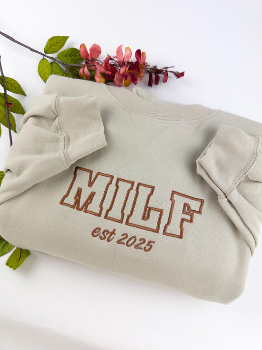 Beige or sand colored sweatshirt with the word Milf written in large brown letters. Under the word Milf are the words est 2025.