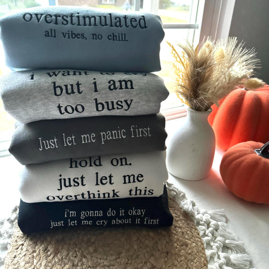 an image of 5 sweatshirt folded on top of one another. Each sweatshirt is a different color and have different text embroidered onto it in various thread colors.