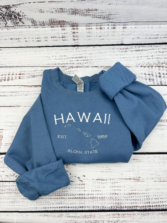 Hawaii embroidered sweatshirt with map and lettering, blue color, folded on white wooden background.