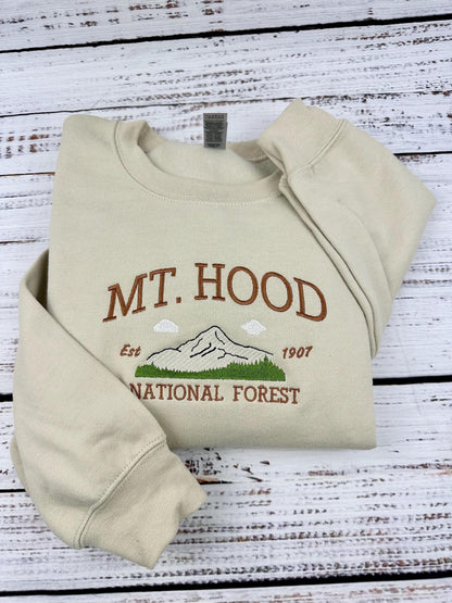 A beige sweatshirt with an embroidered design of Mt. Hood National Forest. The design includes a mountain, clouds, trees, and the text &#39;Mt. Hood&#39;, &#39;Est. 1907&#39;, and &#39;National Forest&#39;.