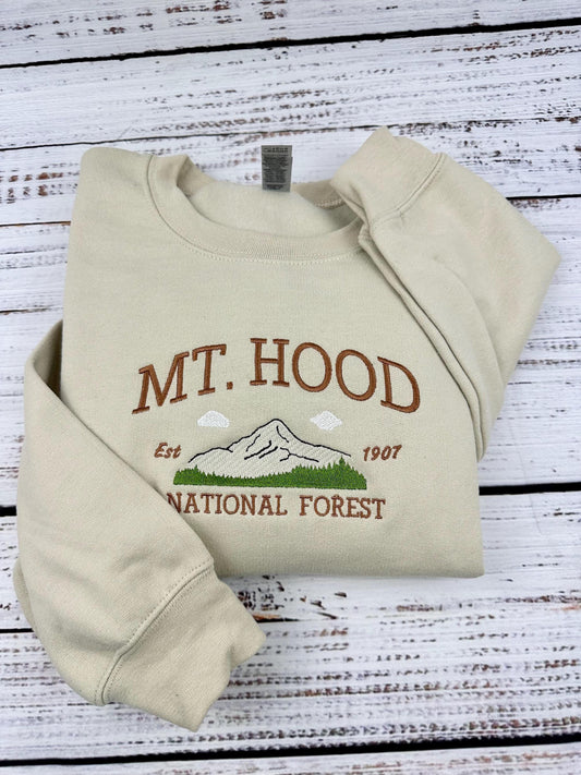 A beige sweatshirt with an embroidered design of Mt. Hood National Forest. The design includes a mountain, clouds, trees, and the text &#39;Mt. Hood&#39;, &#39;Est. 1907&#39;, and &#39;National Forest&#39;.