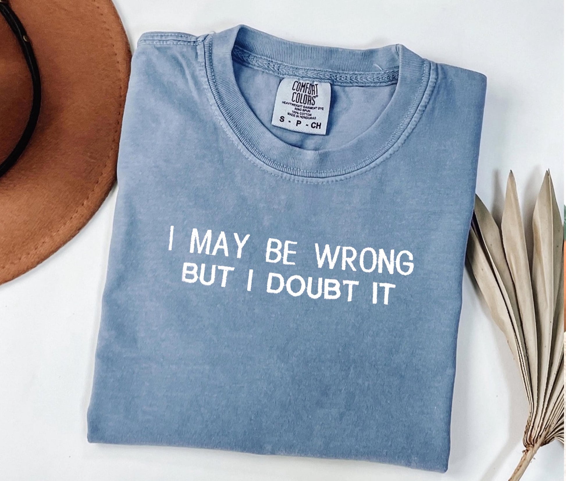 Personalized Comfort Colors® embroidered t-shirt with text "I May Be Wrong But I Doubt It," folded next to a hat and dried leaves.