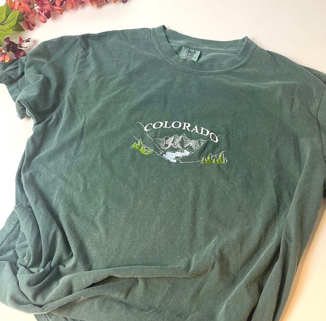 A dark green t-shirt with a white embroidered design of mountains and trees. The design includes the word &#39;COLORADO&#39;.