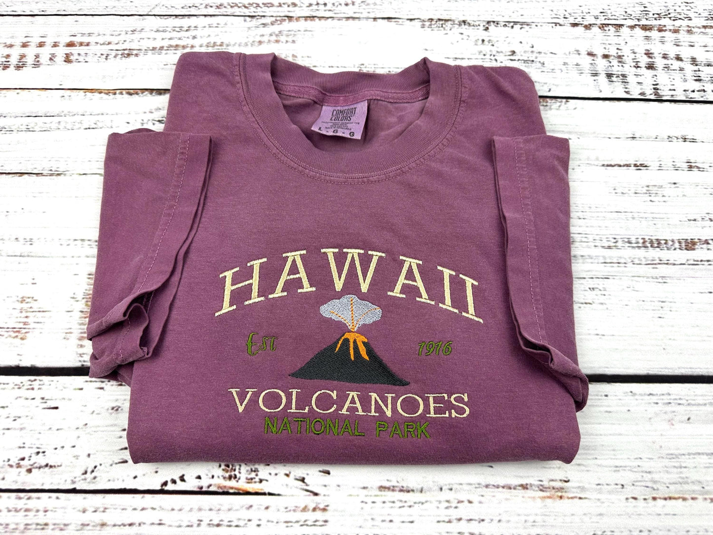 a shirt with an embroidery design of Hawaii volcanoes national park. image of an erupting volcanoe is under the text