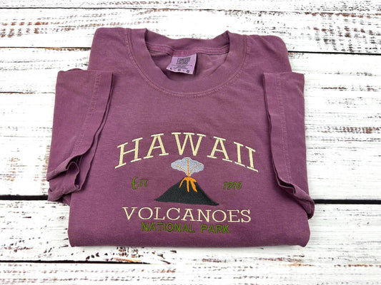 a shirt with an embroidery design of Hawaii volcanoes national park. image of an erupting volcanoe is under the text
