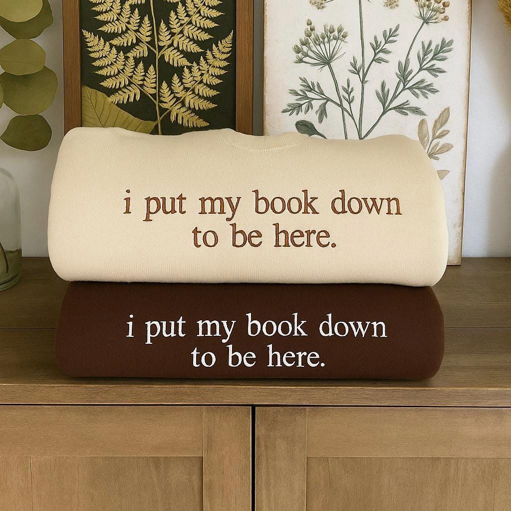 two shirts stacked above one another. the top is sand with brown thread and the bottom is brown with white thread. The text embroidered says I put my book down to be here